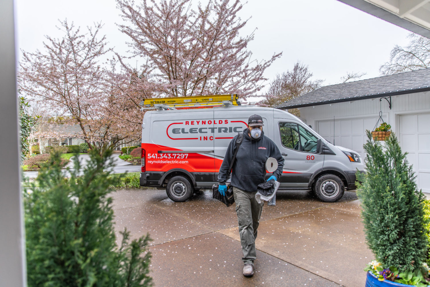 A Reynolds Electric and Plumbing technician walking toward a home with the service van behind him.