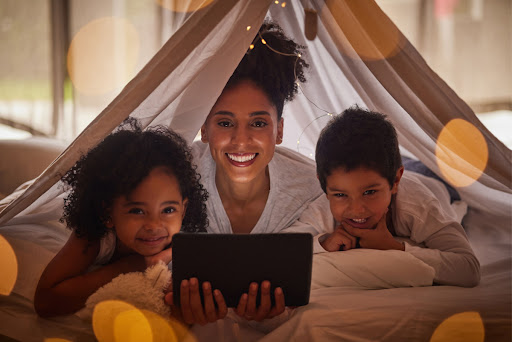 A mom and two kids sitting in a blanket fort with a tablet.