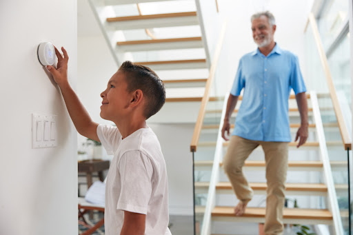 A kid adjusting a thermostat as an older man descends a staircase in the background.